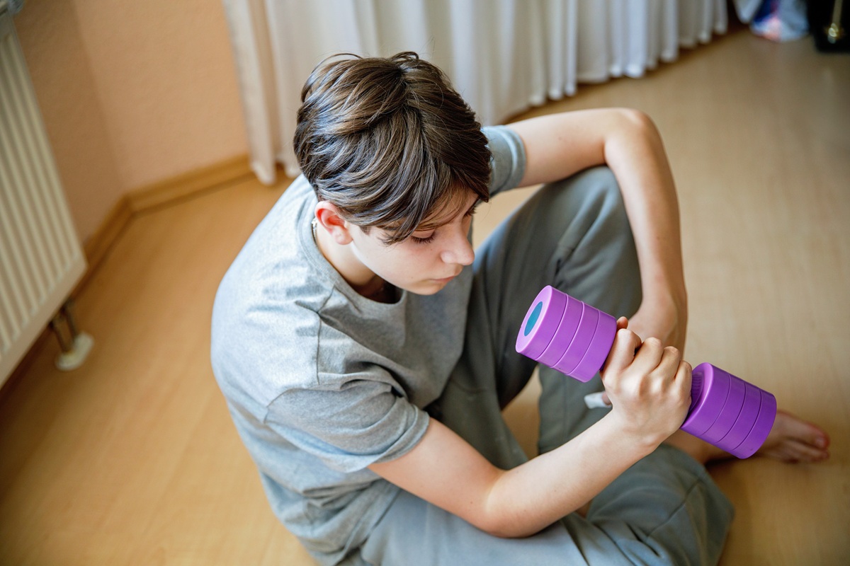 Osoba siedzi na podłodze w pomieszczeniu, ubrana w luźny strój sportowy, trzyma w dłoni fioletowy hantel.